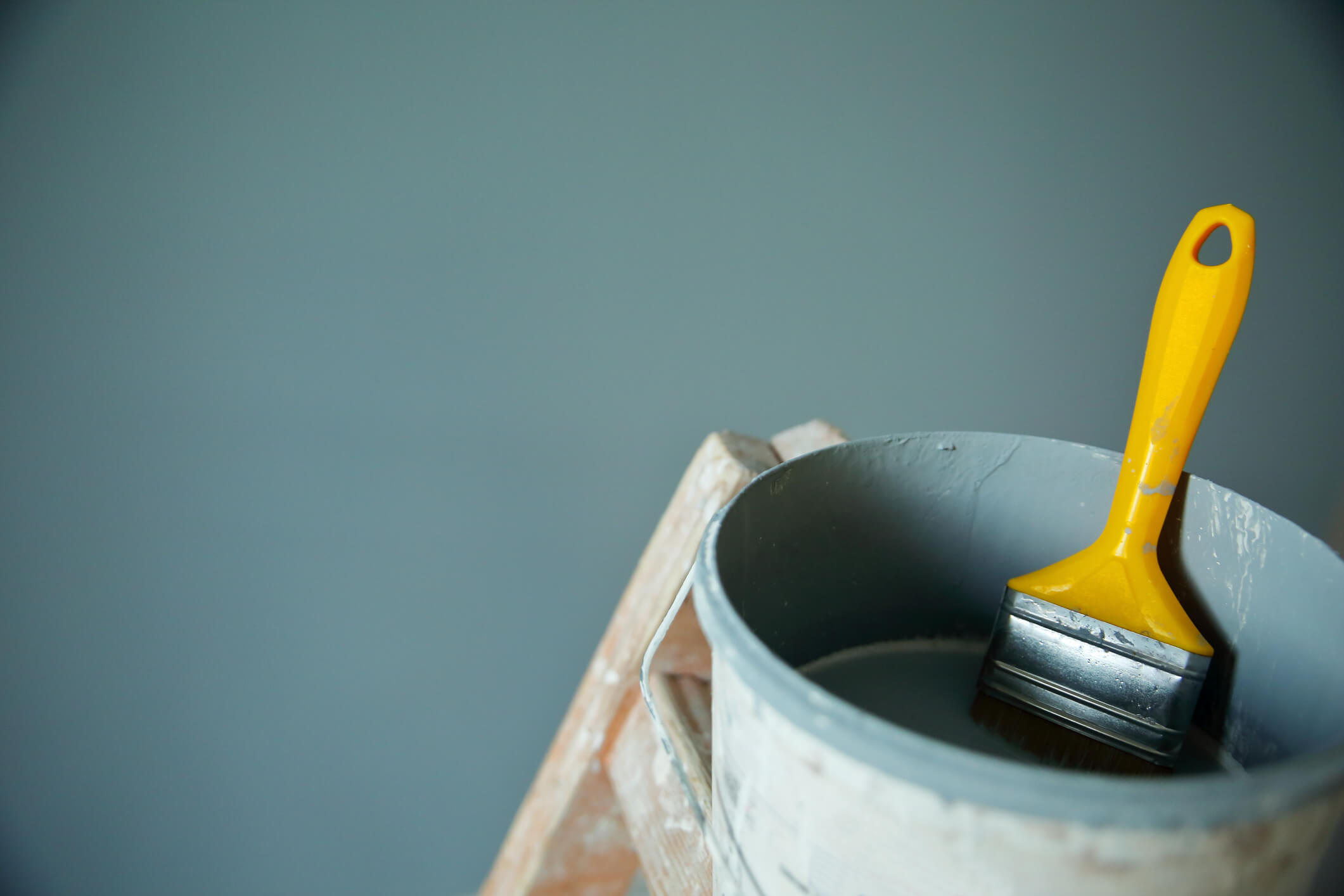 An open paint bucket with a paint brush in front of a freshly painted wall